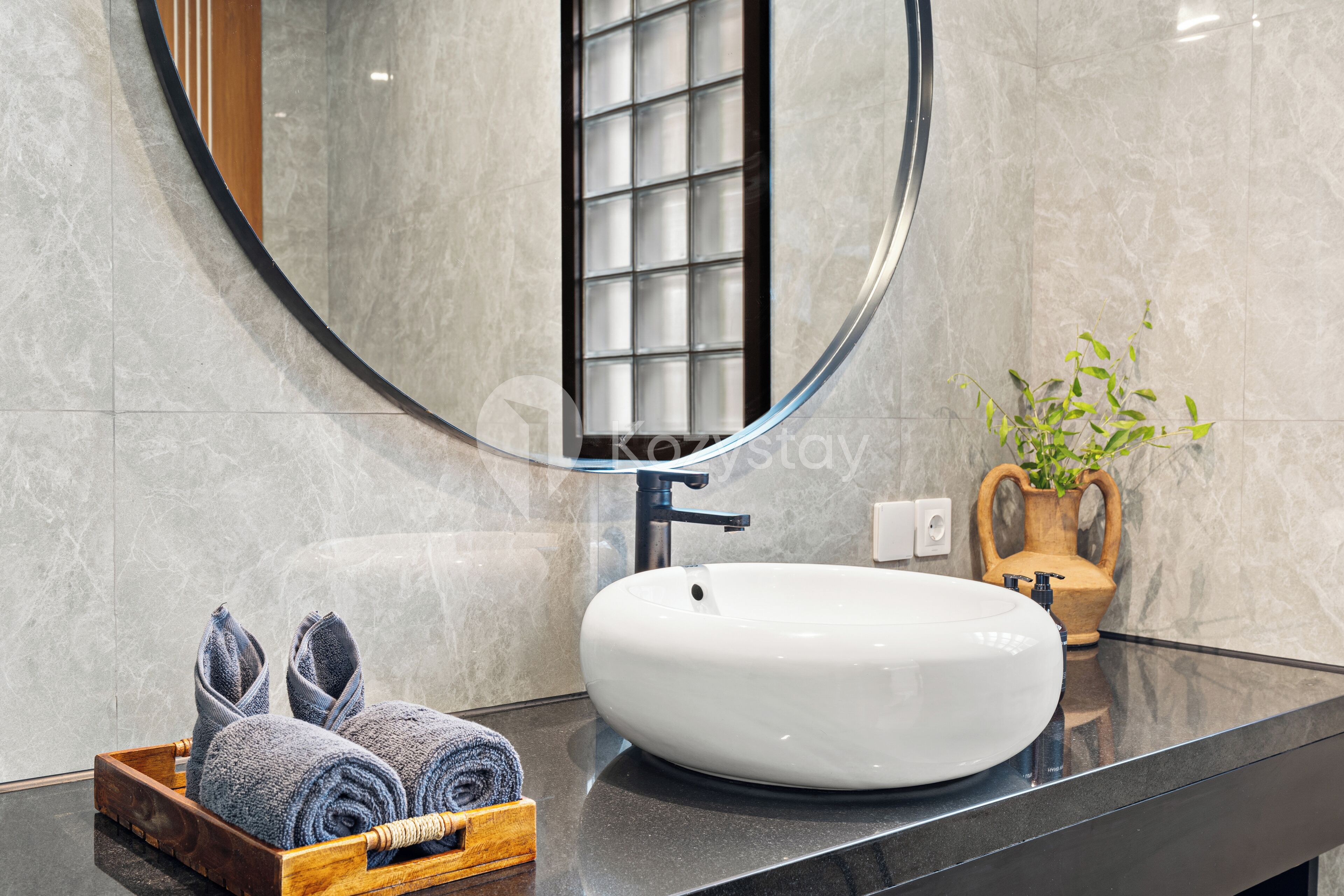 Un baño con un espejo redondo, un lavabo blanco y una bandeja de madera con toallas.