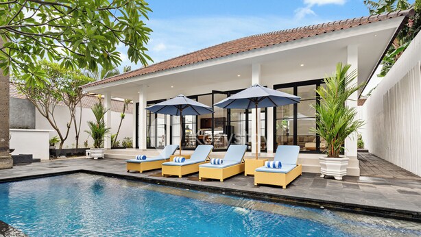 A pool area with lounge chairs and umbrellas, surrounded by a white building with a tiled roof. - Kros Villa by Kozystay - Legian (Kuta)