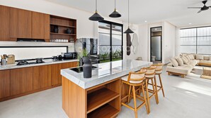 A modern kitchen with wooden cabinets, a marble countertop, and a central island with bar stools. - Kros Villa by Kozystay - Legian (Kuta)