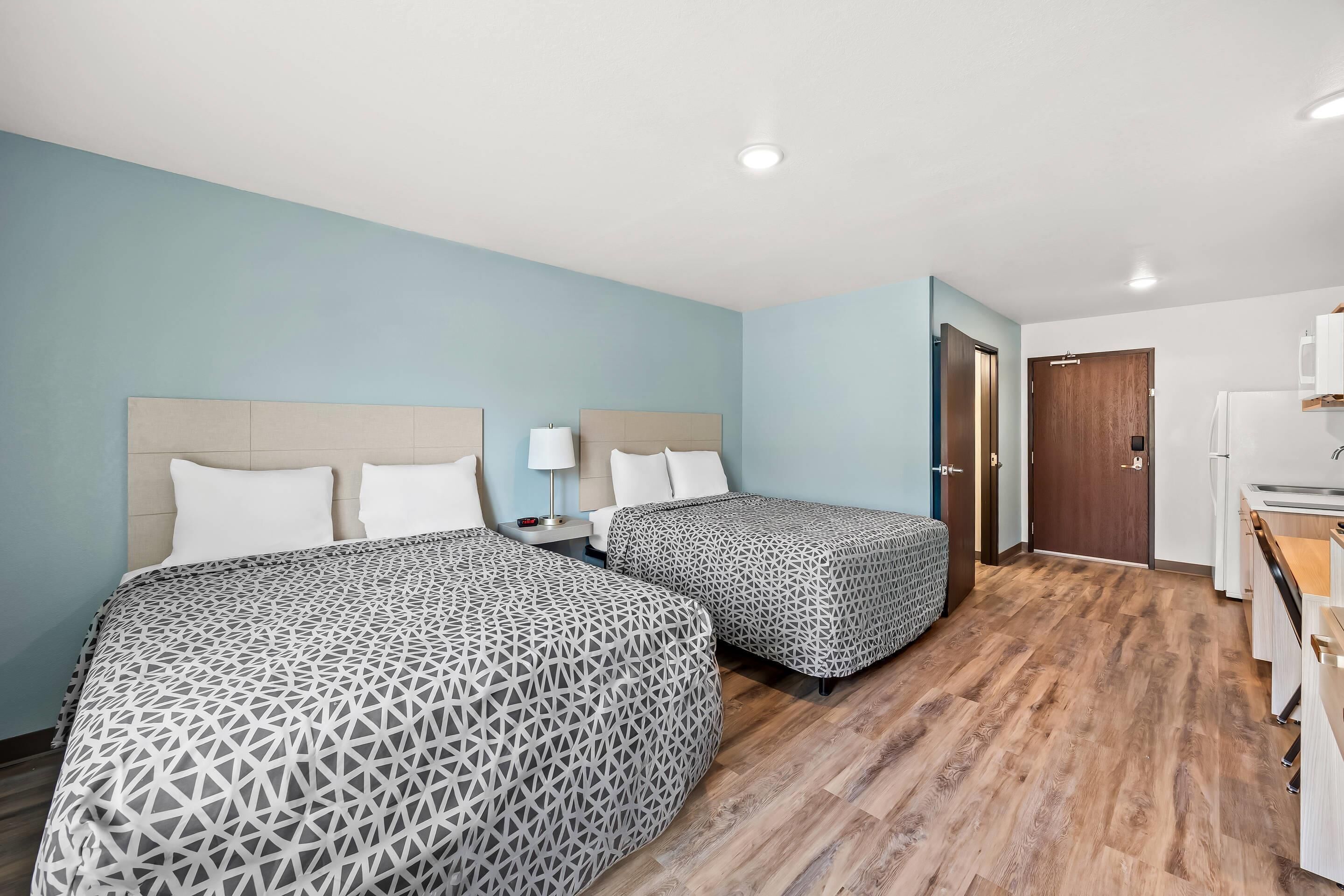 A hotel room with two beds, a wooden floor, a kitchenette, and a door leading to another room.