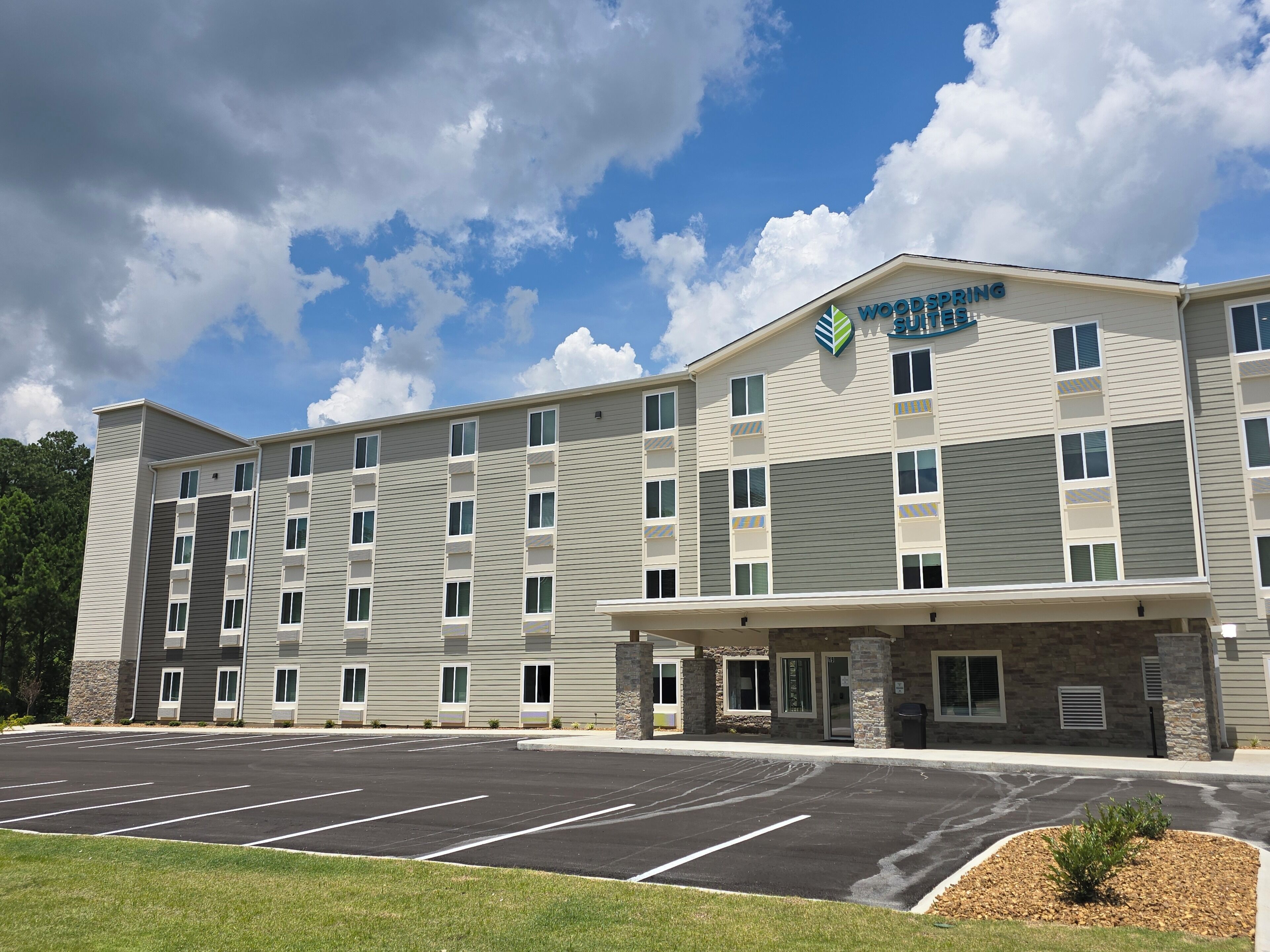 A multi-story hotel with a parking lot in front, featuring a modern design with a mix of grey and beige exterior walls and stone accents.