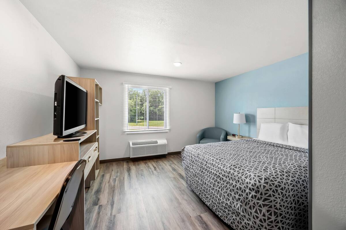 A modern hotel room with a wooden desk, a flat-screen TV, a bed with a patterned bedspread, a window with blinds, and a nightstand with a lamp.