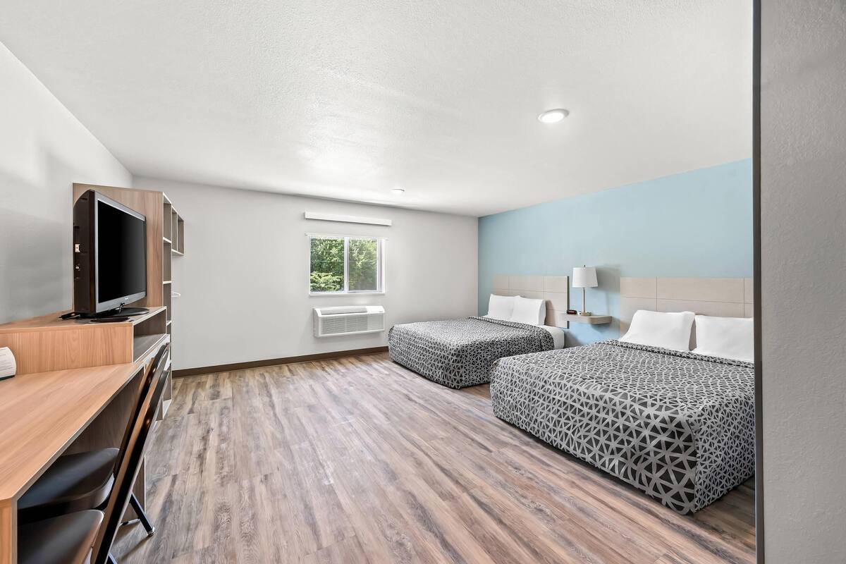 A hotel room with a wooden desk, a television, two beds with patterned bedding, a window, and a ceiling light.