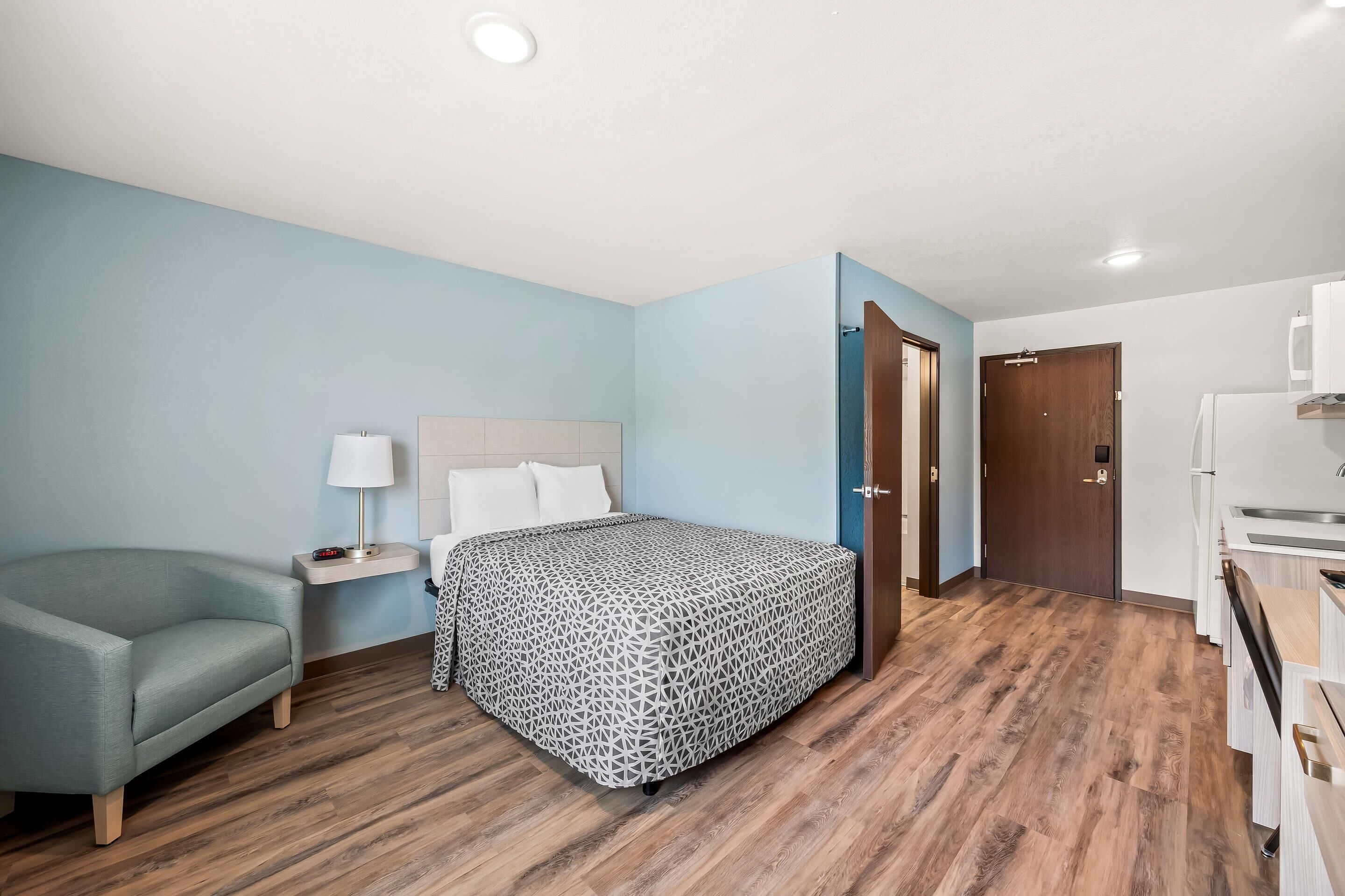 A hotel room with a bed, a nightstand, a lamp, a sofa, a wooden floor, a door, and a kitchenette.