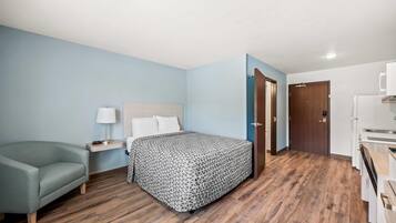 A hotel room with a bed, a nightstand, a lamp, a sofa, a wooden floor, a door, and a kitchenette.