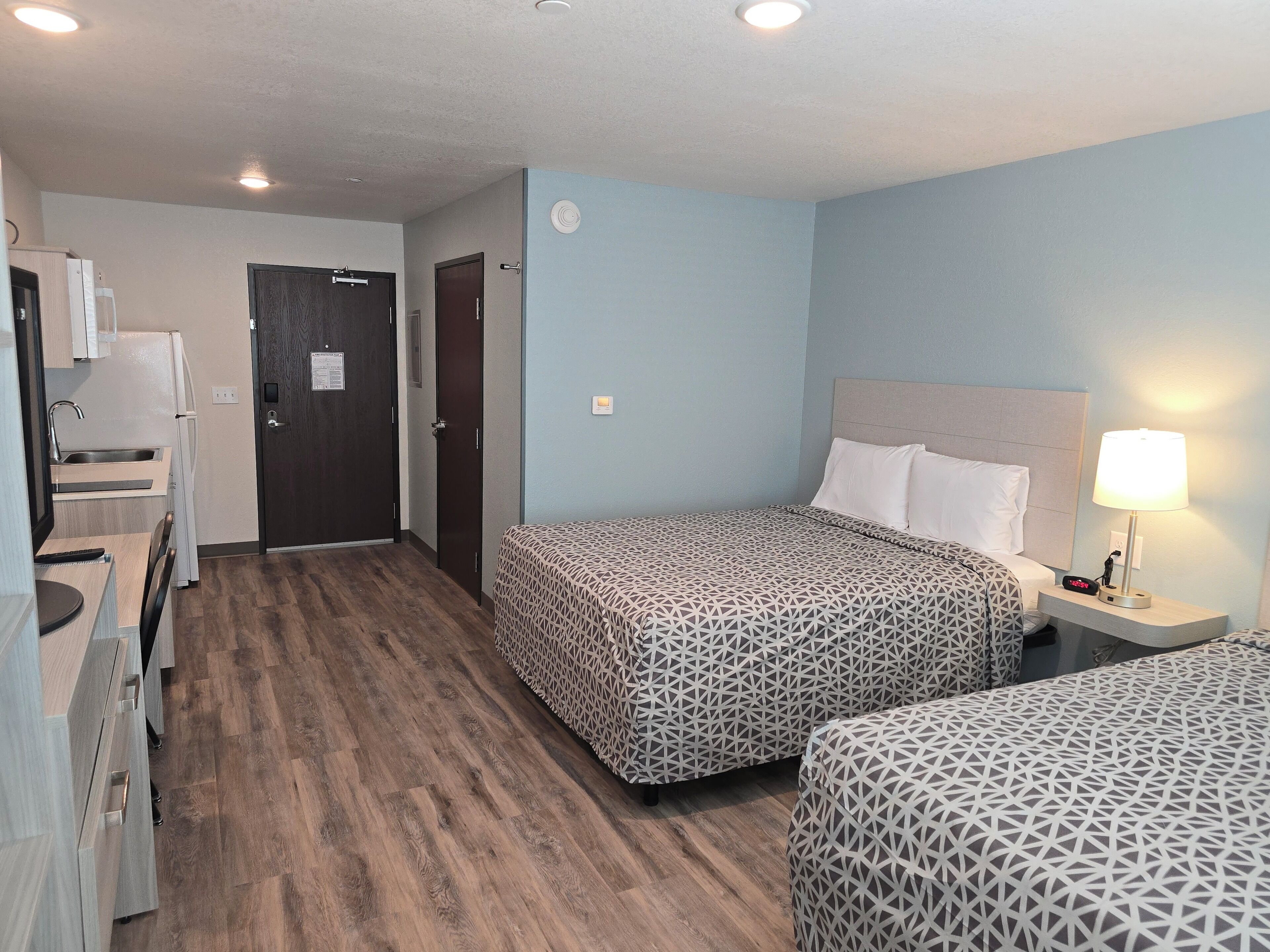 Habitación de hotel con dos camas, un escritorio con silla, una kitchenette pequeña y la puerta del baño.