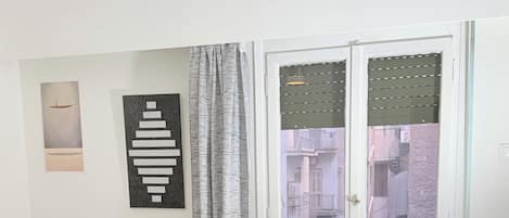 A bedroom with a bed, a chair, a sofa, a hanging light, a window with blinds, and an air conditioning unit.
