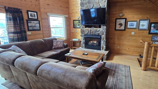 A cozy cabin interior with a stone fireplace, a large flat-screen TV mounted above it, a brown leather sofa, and a wooden coffee table. - Spacious Wisp Retreat w/ Views & Game Room (Mchenry)