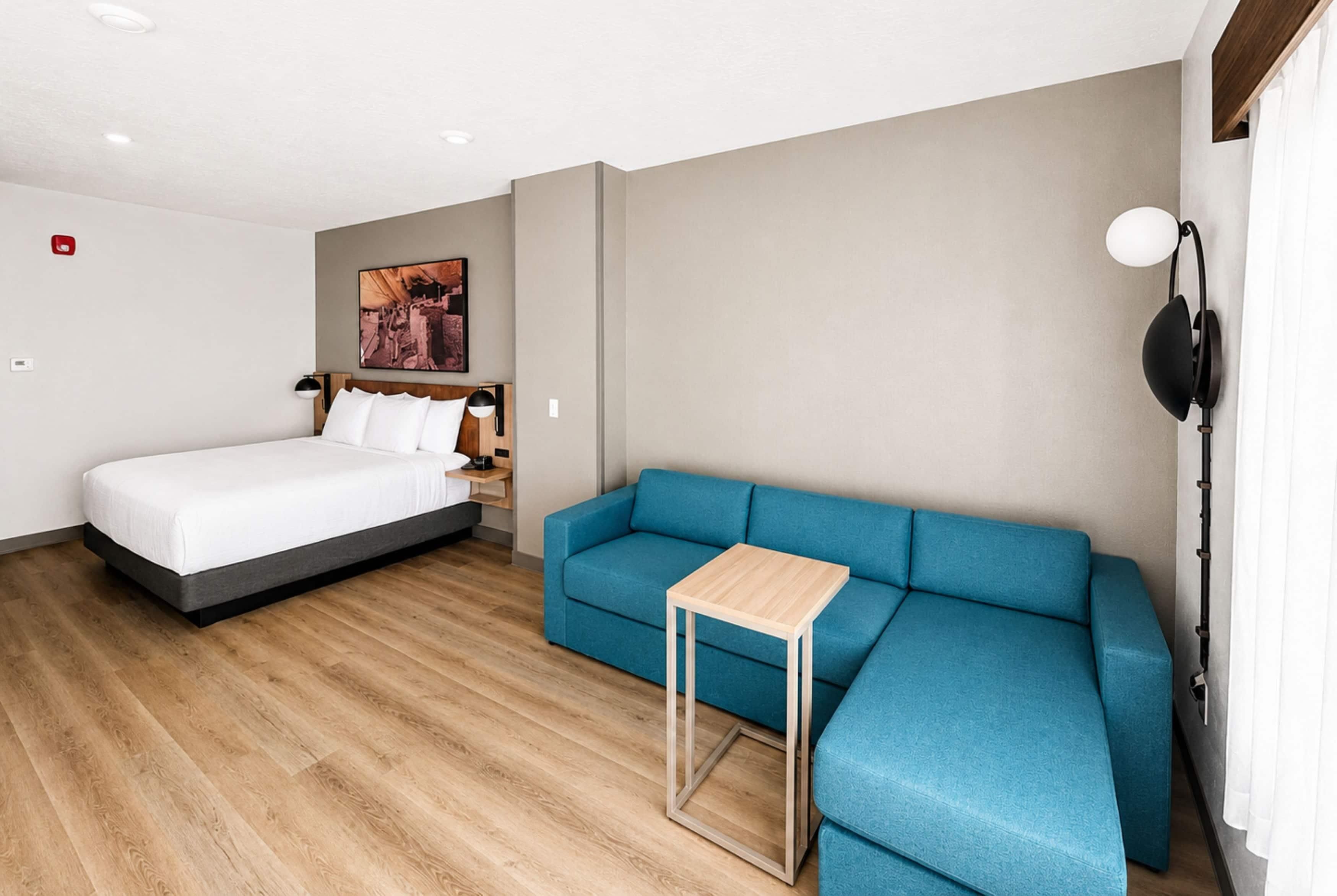 A modern hotel room with a bed, a blue sofa, a small table, a wall-mounted lamp, and a painting.