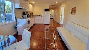 Una cocina compacta con armarios blancos, estufa, microondas y refrigerador. Hay una mesa de comedor de vidrio y un sofá blanco en la sala.