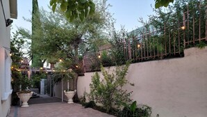 Un patio empedrado con suelo de baldosas, una pared blanca y una valla de madera. Hay plantas en macetas y una guirnalda de luces.