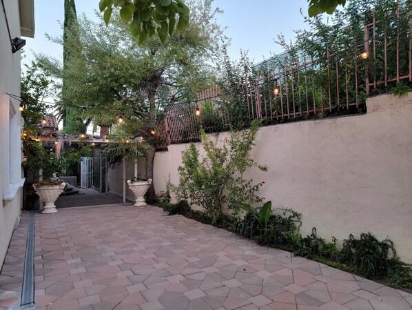A paved courtyard with a tiled floor, a white wall, and a wooden fence. There are potted plants and a string of lights.
