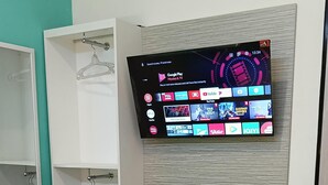 A hotel room with a bed, a TV mounted on the wall displaying a streaming service interface, a wardrobe with hangers, and a small shelf. - Comfort Centre Residence Solo (Surakarta)