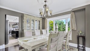 Une salle à manger avec une grande table, des chaises, un lustre et une vue sur l’extérieur.