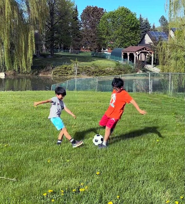 Two children playing soccer on a grassy field with a pond and trees in the background.