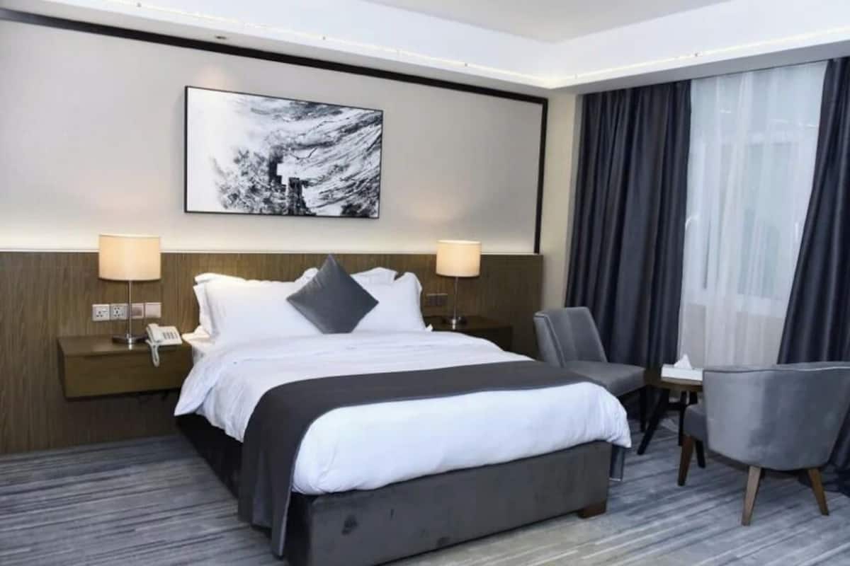Hotellihuone, jossa on sänky, kaksi yöpöytälamppua, tuoli, pieni pöytä ja taulu seinällä.