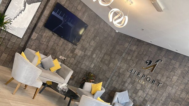 Una hall d'albergo moderna con un'area salotto che presenta divani grigio chiaro con cuscini gialli, un tavolino rotondo e una TV a schermo piatto montata a parete. Il pavimento è piastrellato e le pareti hanno una superficie strutturata.