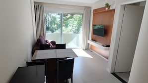 Ein modernes Wohnzimmer mit Essbereich, einem an einer Holzplatte montierten Fernseher und einem Balkon mit Blick auf Grünflächen.