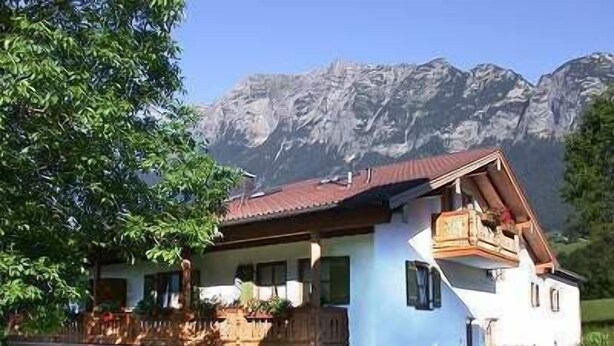 A two-story chalet with a balcony, surrounded by greenery and mountains.