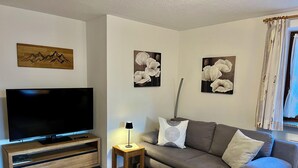 A living room with a flat-screen TV on a wooden stand, a grey sofa with cushions, and two wall-mounted flower paintings.