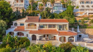 Una villa de varios pisos con techo de tejas, rodeada de abundante vegetación y otras edificaciones residenciales.
