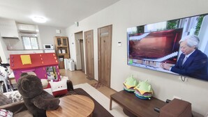Un salon avec un téléviseur diffusant un programme d’information, une table en bois, un canapé et un parc pour enfants avec un ours en peluche.