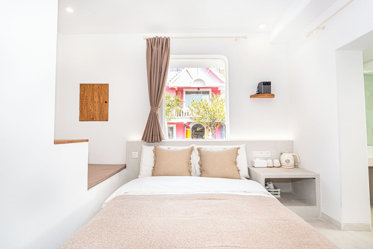 A modern bedroom with a large bed, a window with curtains, a bedside table with a kettle, and a small shelf.