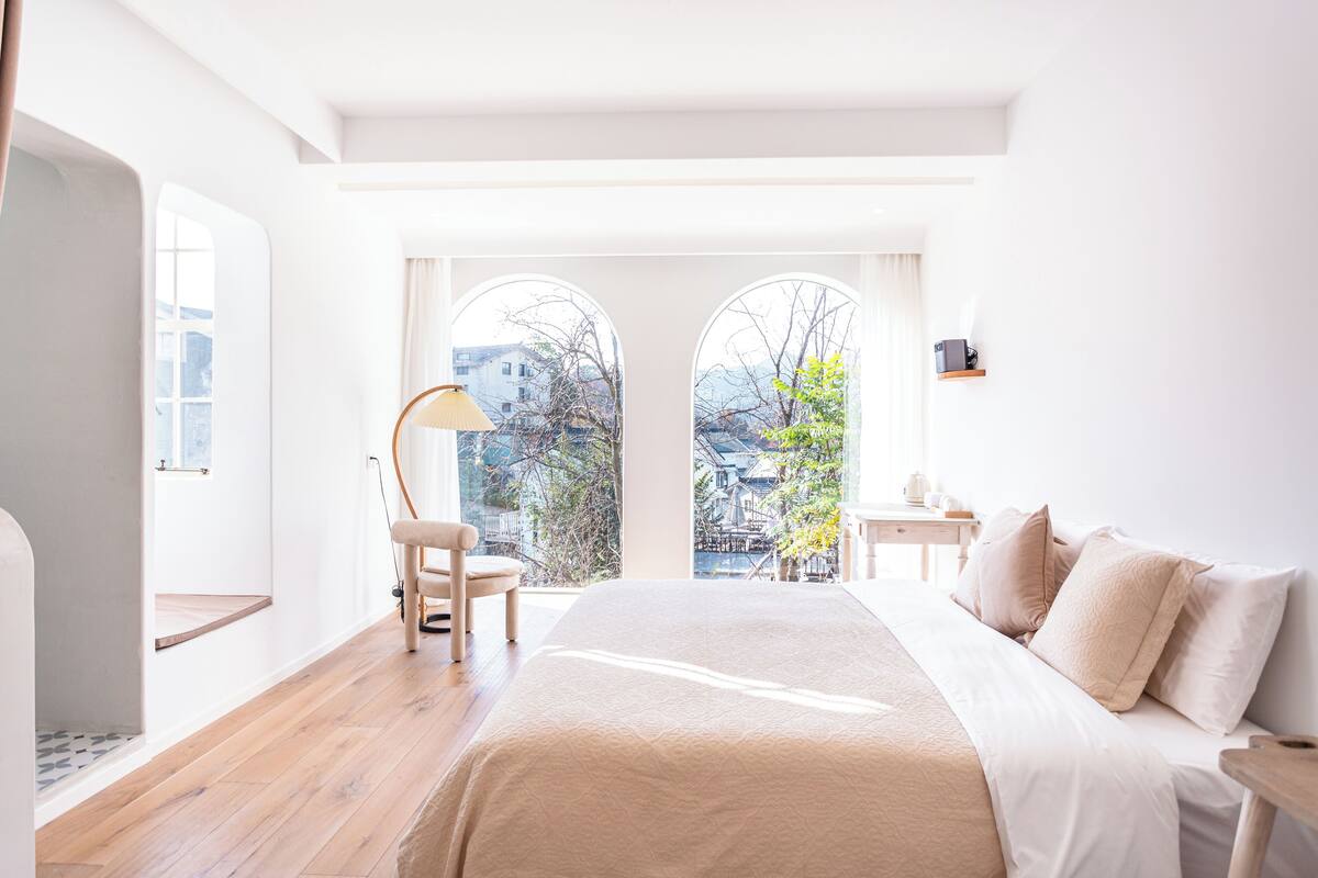 A bedroom with a large bed, a chair, a nightstand, and a view of a building through a window.