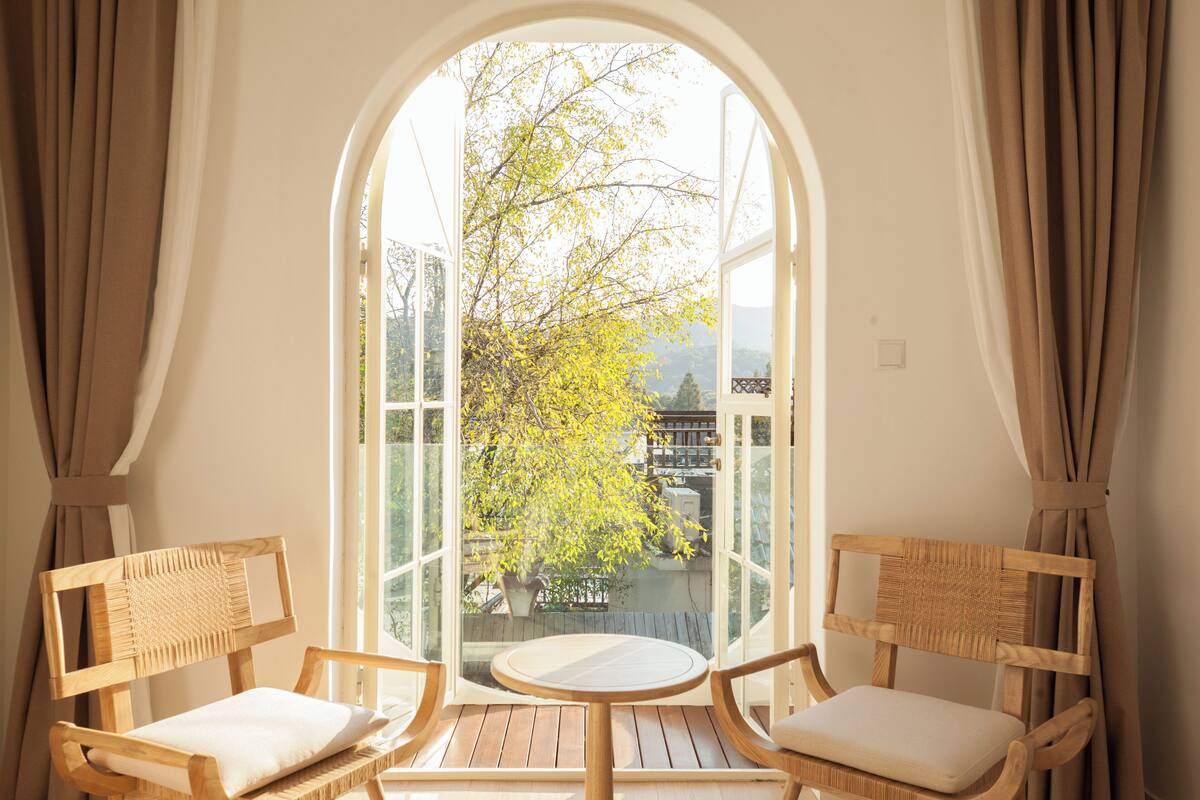 A room with two wooden chairs and a small round table, a large arched window, and a view of trees and buildings outside.
