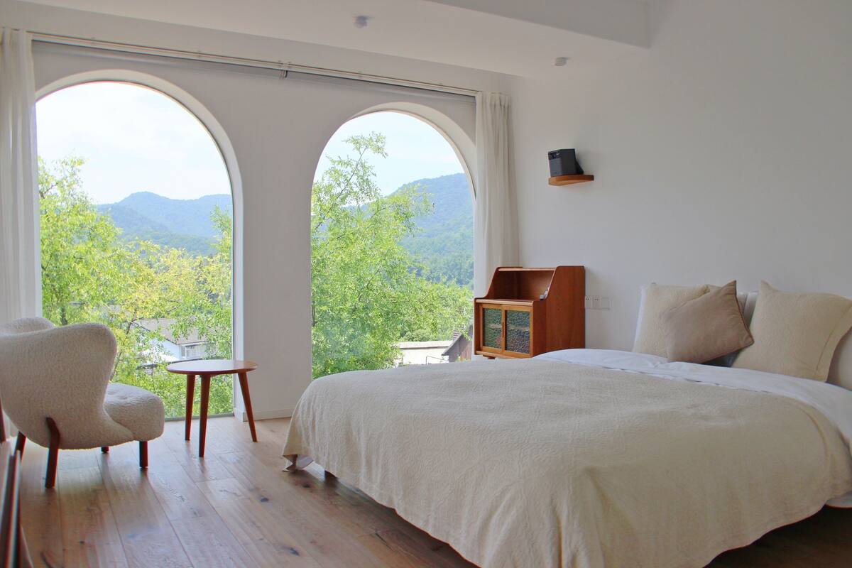 A bedroom with a large bed, a chair, and a small table. There are two large windows with a view of trees and mountains.