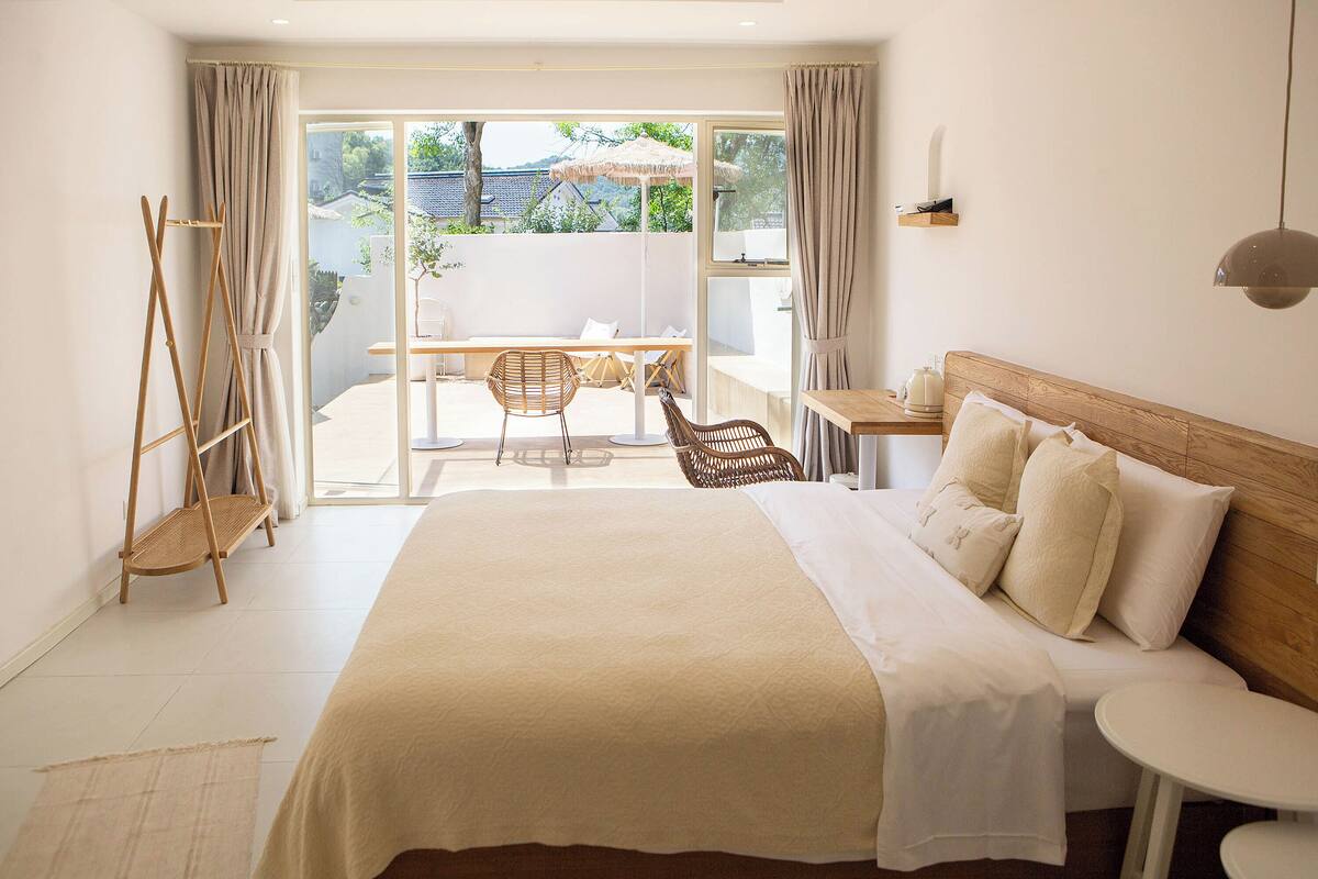 A bedroom with a large bed, a small table, a chair, and a view of the outdoors.