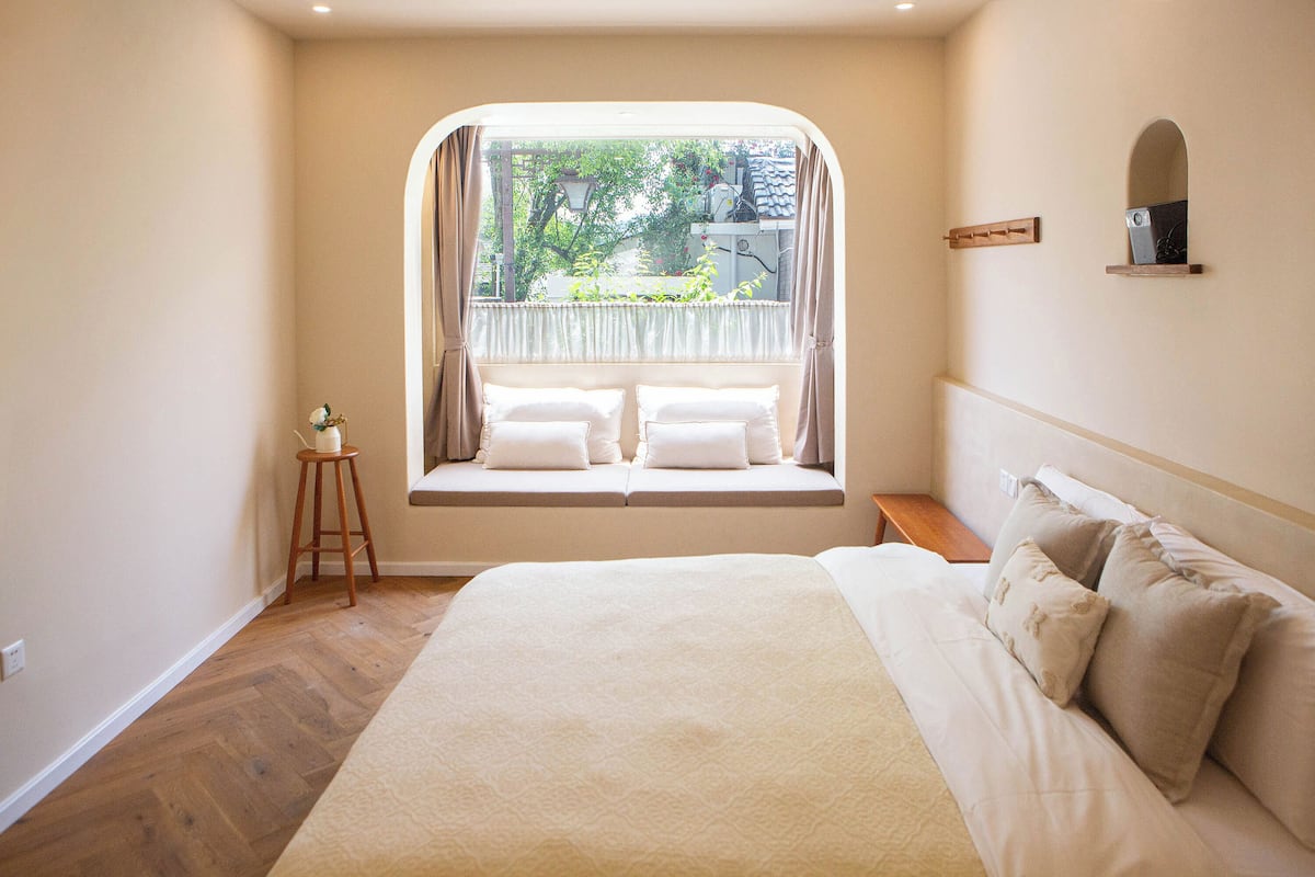 A bedroom with a large bed, a window seat with cushions, a small stool, and a bedside table.