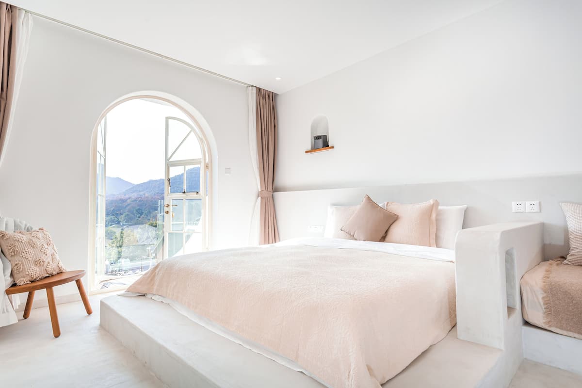 A bedroom with a large bed, a small seating area, a window with a view of mountains, and a shelf with a lamp.