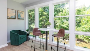 A room with a green sofa, two bar stools, a round table, and large windows with a view of trees. - Zinnia- Deluxe Double Queen Studio at The Dogwood (Maggie Valley)