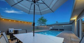 Un área junto a la piscina con una mesa, sillas, una sombrilla, luces colgantes y una parrilla.