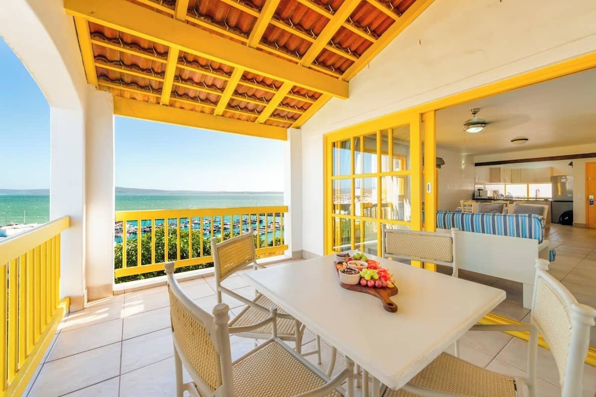 A balcony with a view of the sea, a dining area with a table set for two, and a kitchen visible through an open door.