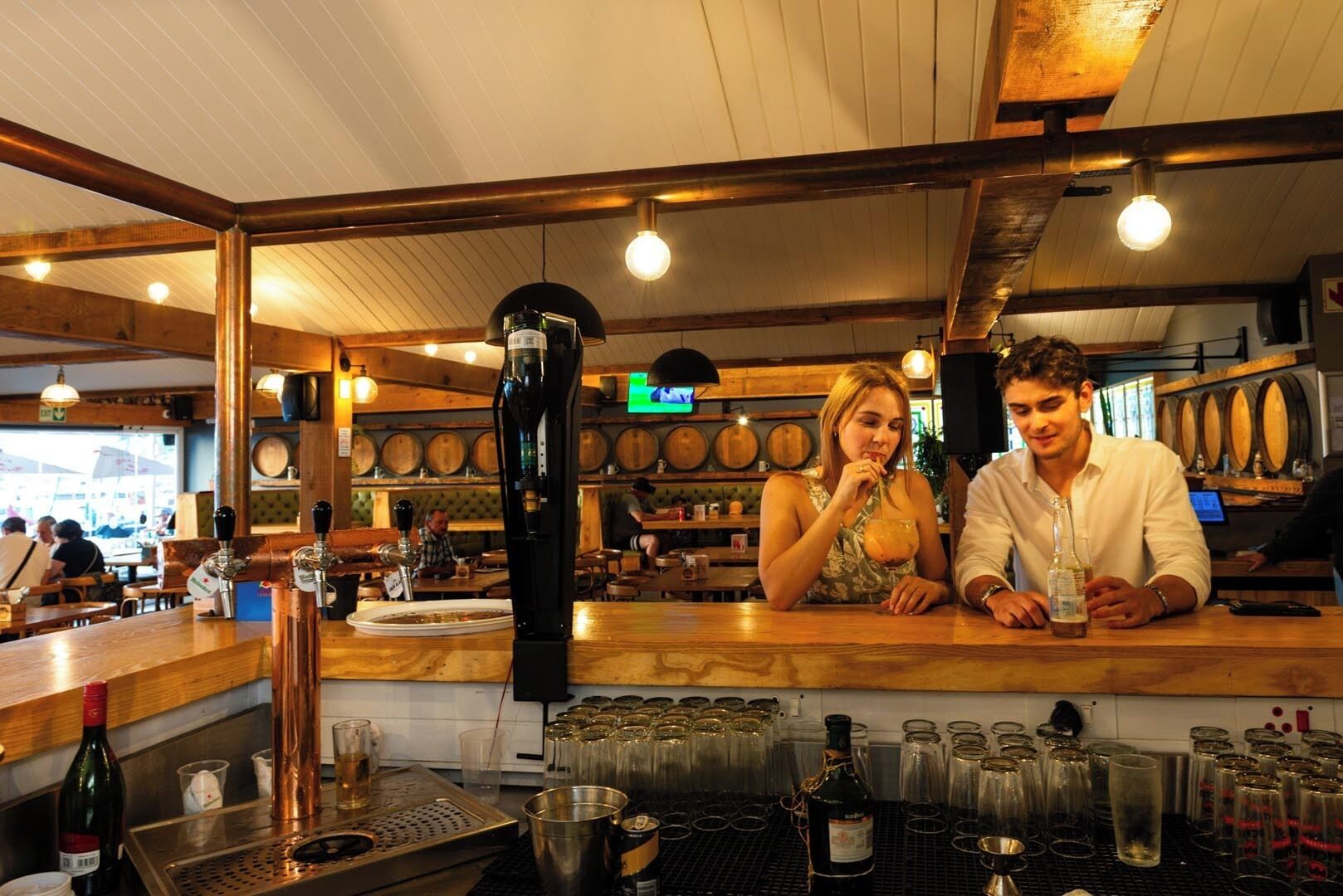 A bar area with a bartender, a woman and a man sitting at the bar, and a counter with various bottles and glasses.