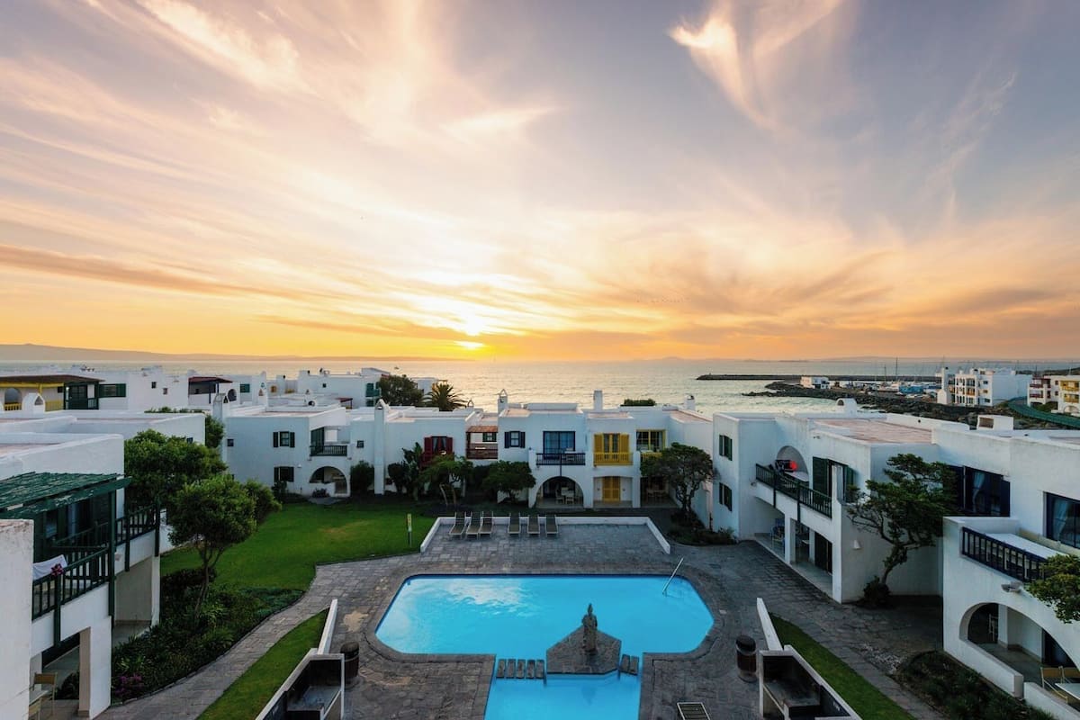 A resort with white buildings, a swimming pool, and a view of the sea at sunset.