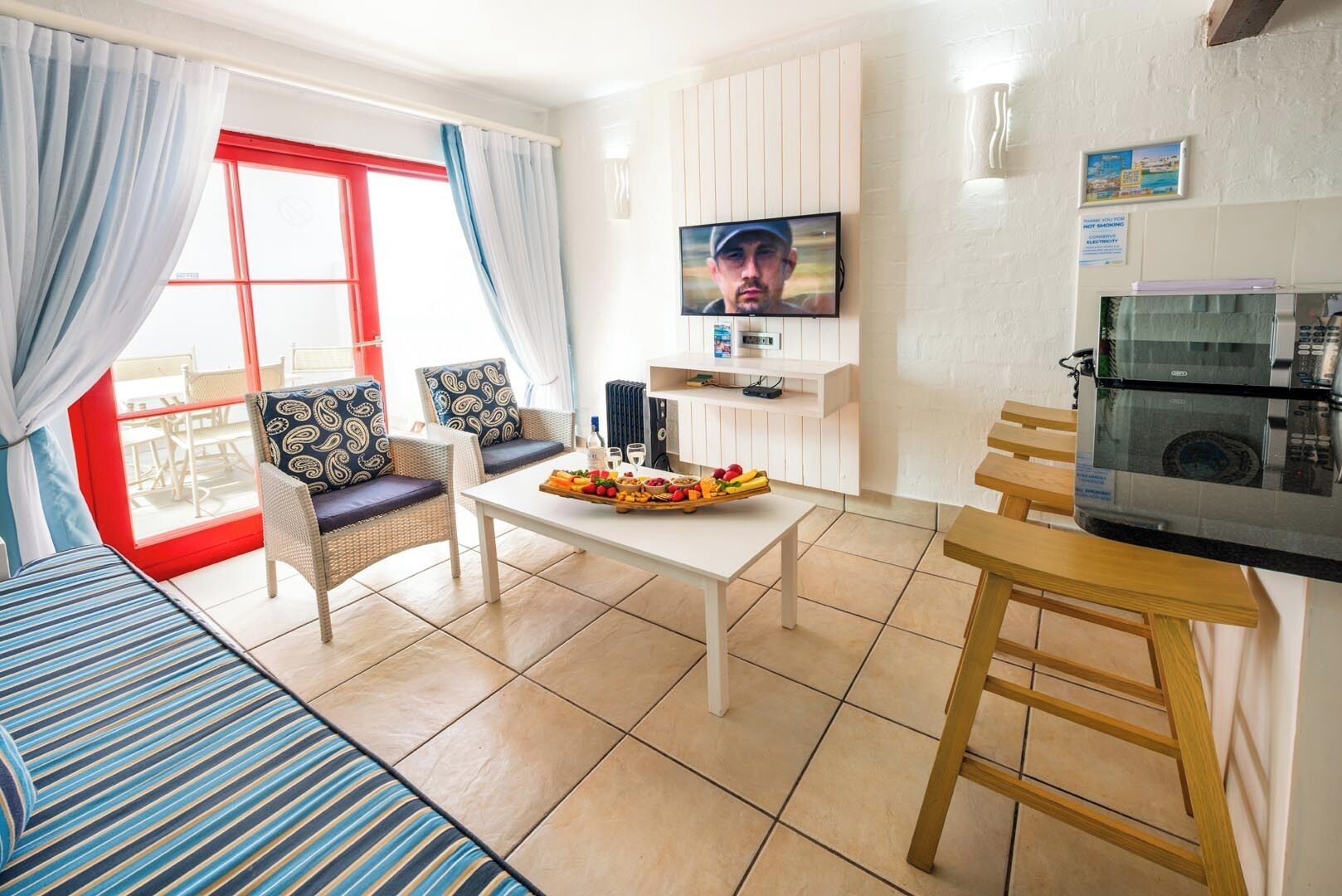 A modern hotel room with a sofa, dining table, and chairs. A TV is mounted on the wall, and there is a kitchen area with a microwave.