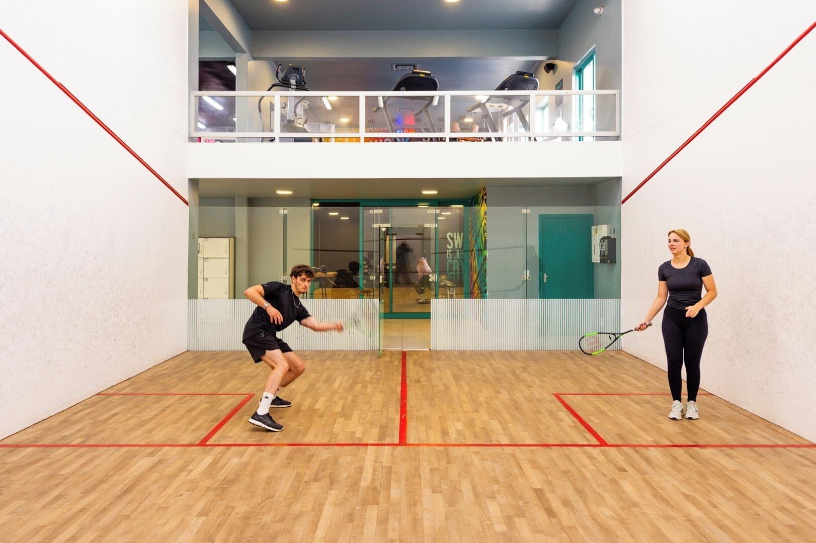En squashbane med trægulv, glasvæg og en dør til et rum med træningsudstyr.