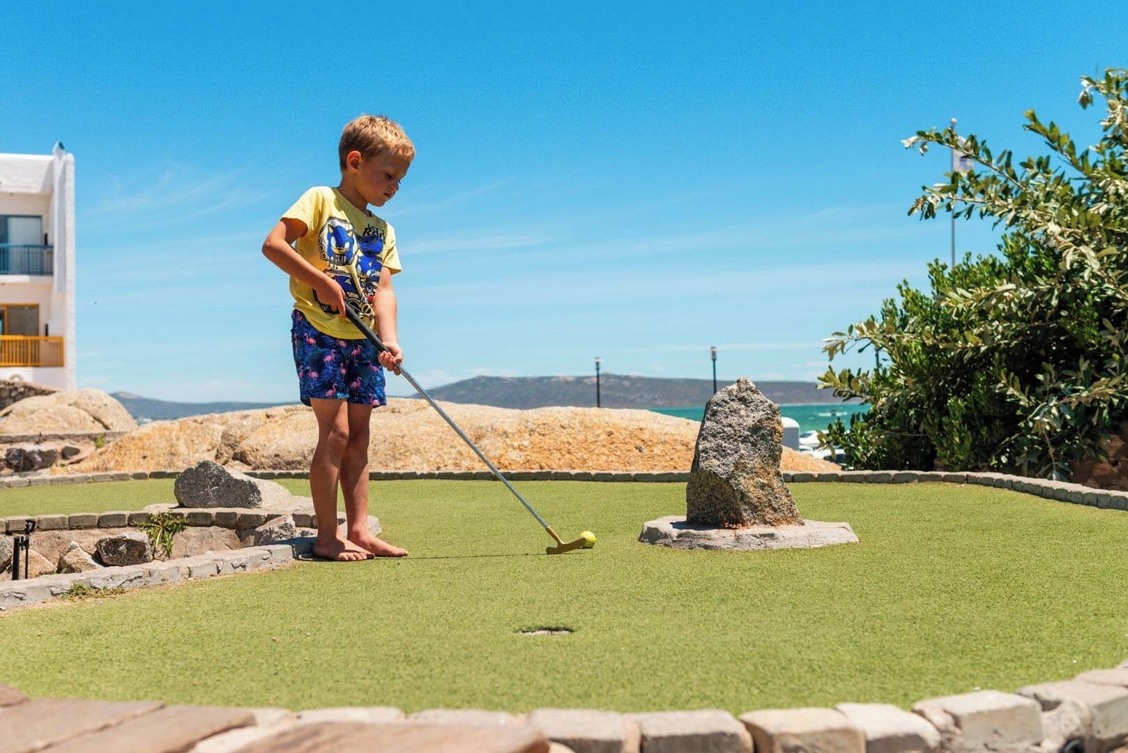 A child playing mini-golf on a green artificial turf with a rocky backdrop and a building to the left.