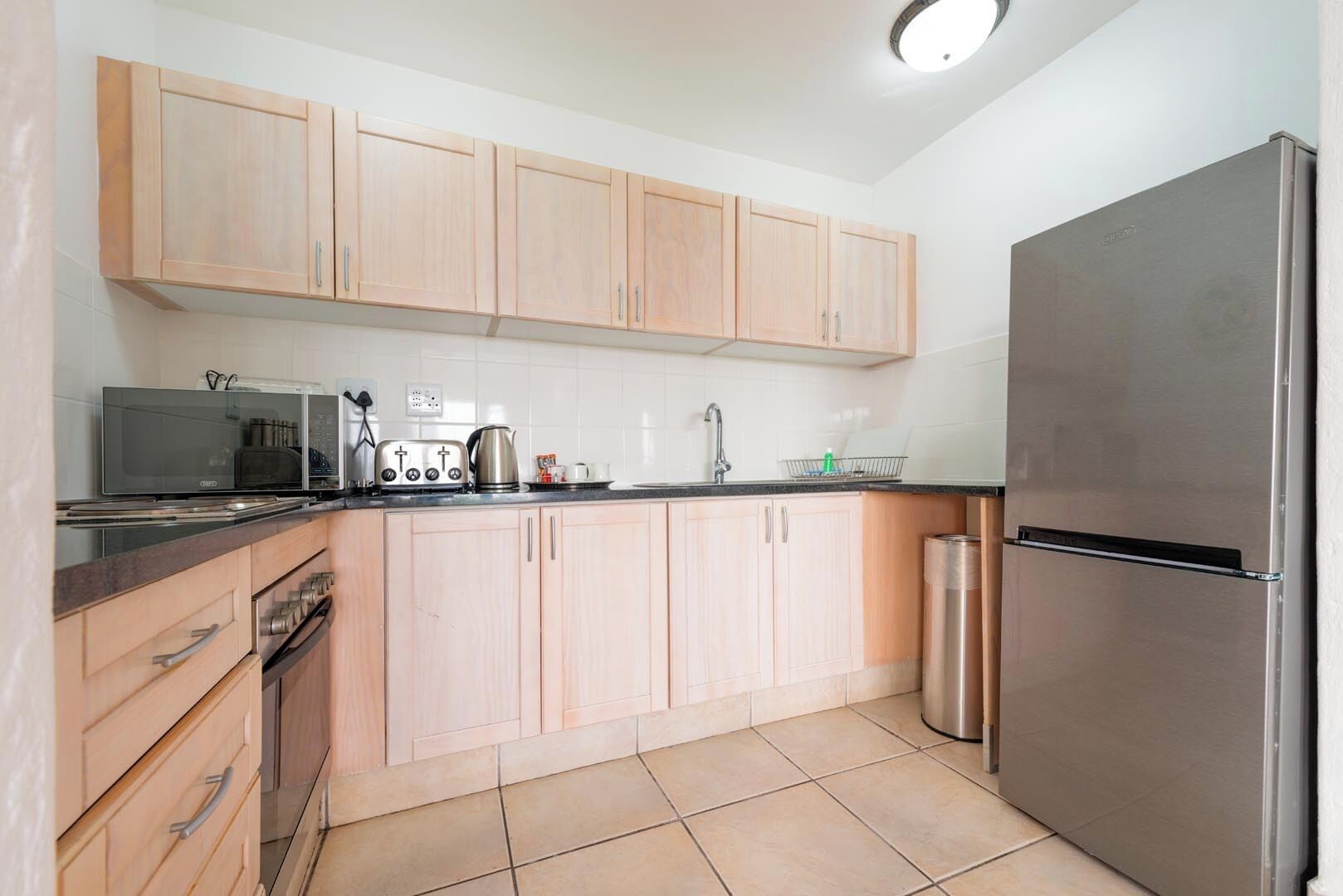 A kitchen with light wooden cabinets, a stainless steel refrigerator, a microwave, a toaster, and a kettle.