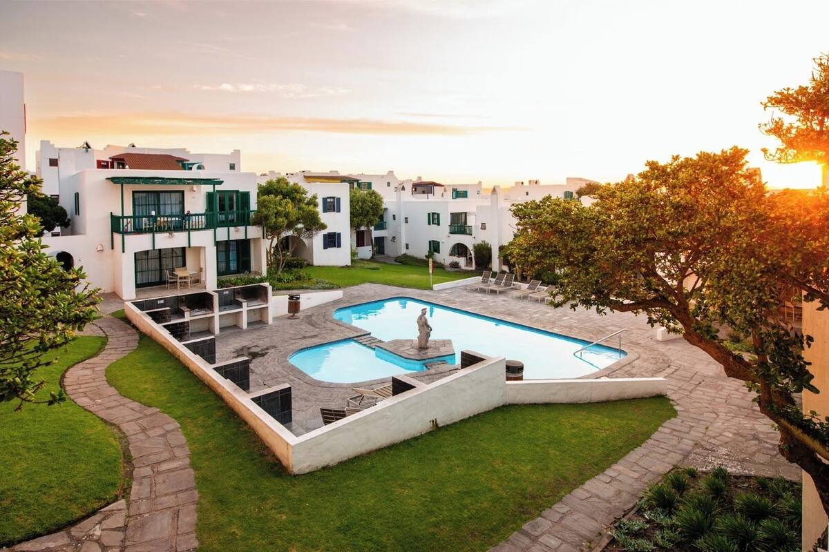A pool area with a statue, surrounded by a white building with balconies and a garden.