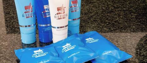 Un set de productos de cuidado corporal Club Mykonos que incluye gel de ducha, loción corporal y champú, junto con jabón de baño y paquetes de gorro de ducha.