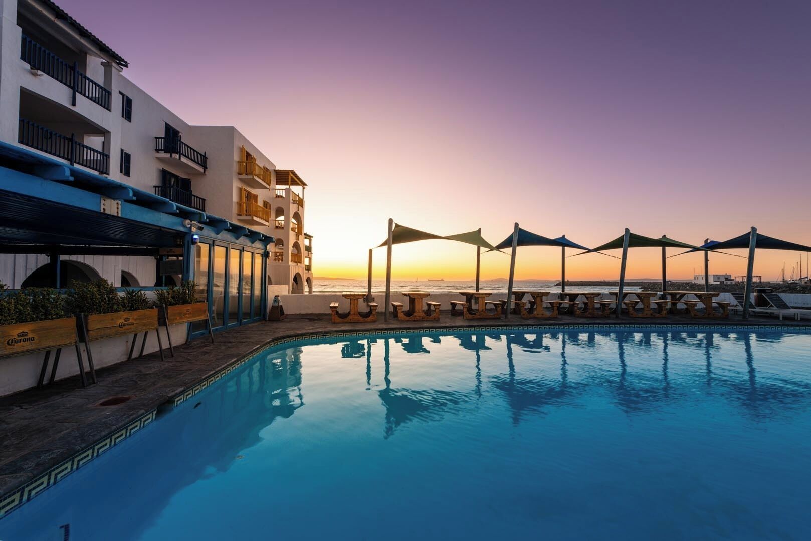 Zona junto a la piscina con vista al atardecer, mobiliario exterior y un edificio con balcones.