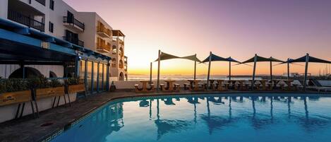 Zona junto a la piscina con vista al atardecer, mobiliario exterior y un edificio con balcones.