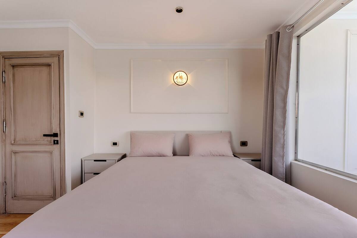 Ein Schlafzimmer mit einem großen Bett, einem Nachttisch, einer Kommode, einer Tür und einem Fenster mit Vorhängen.