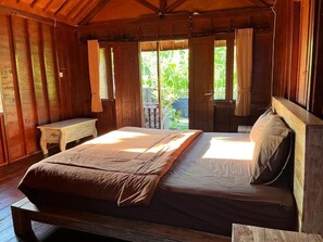 A wooden room with a large bed, a small table, and a view of greenery outside. - Telagamas Guest House (Banjarangkan)