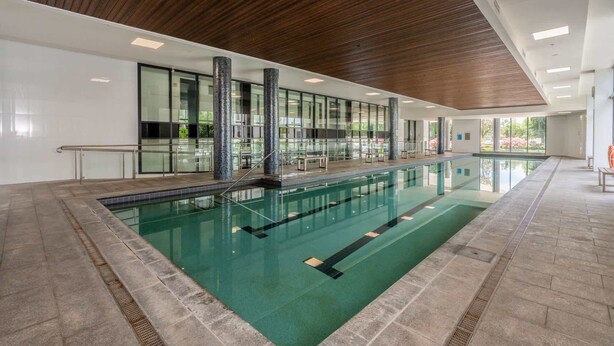 Une piscine intérieure moderne, avec de l’eau cristalline, entourée de murs en verre et surmontée de poutres en bois.
