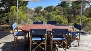 A wooden deck with a round table and six chairs, two large umbrellas, and a backdrop of trees and shrubs. - 15% Off Weekly | Coastal-View Cypress Beach House (Island Beach)
